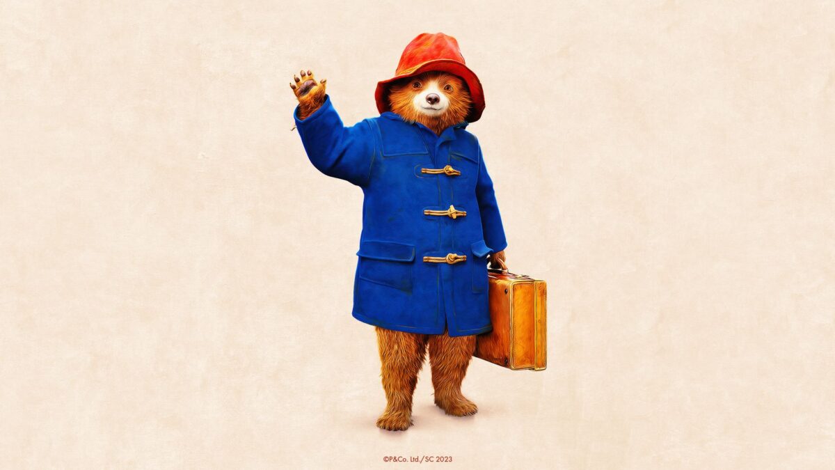 Image of Paddington Bear waving at the camera. He is wearing a blue raincoat and red hat with a suitcase in his other hand