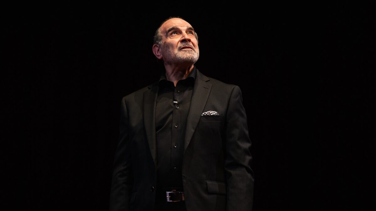 David Suchet in a black suit standing on a black stage, looking up off camera